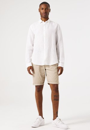 Witte linnen overhemd met lange mouwen, beige opgerolde shorts, witte sneakers. Man staat met ontspannen armen, eenvoudig ontwerp en neutrale kleuren.