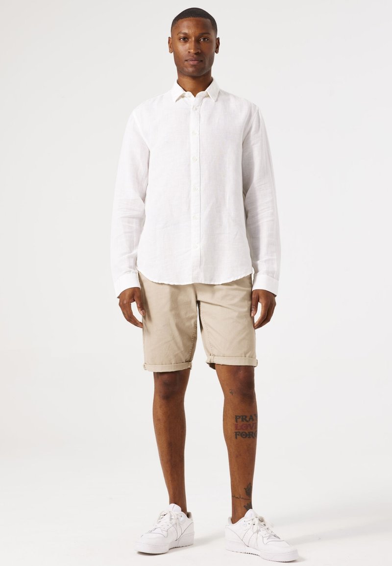 Chemise blanche en lin à manches longues, shorts beige retroussés, baskets blanches. Homme debout, les bras détendus, design simple et couleurs neutres.