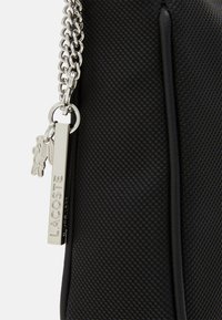 Bolso negro texturizado con una correa de cadena metálica. Presenta un emblema de Lacoste en plata y un detalle de llavero. Diseño rectangular compacto.