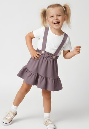 Szczęśliwa młoda dziewczyna o blond włosach w kucykach, ubrana w białą koszulkę i purpurową spódnicę na szelkach, pozująca z jedną ręką na biodrze i jedną uniesioną nogą.