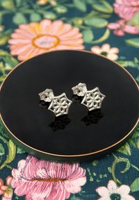 Silverörhängen med geometrisk blommönster, visade på en rund, glansig svart tallrik mot en blommig bakgrund.