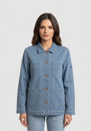 Donna che indossa una giacca di jeans azzurro chiaro con bottoni e quattro tasche frontali, in piedi davanti a uno sfondo grigio chiaro.