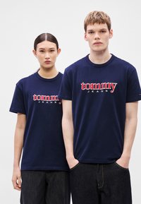 Camisetas de algodón azul marino con el logo "tommy" en rojo y blanco, de manga corta y cuello redondo, combinadas con pantalones vaqueros oscuros.