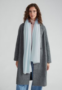 Cappotto grigio in lana con forma oversize, due bottoni e vestibilità rilassata. Scarf leggero blu chiaro che si drappeggia elegantemente attorno al collo. Texture morbida.