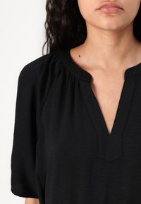 Μαύρη μπλούζα με V-απο neckline, πτυχώσεις γύρω από τον γιακά και κοντά μανίκια. Το ύφασμα έχει ελαφρώς υφασμένο φινίρισμα.