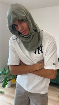 Haină verde cu glugă și șnururi, purtată peste un tricou alb cu un logo negru. Material ușor, pliat, cu brațele încrucișate.