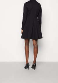 Kvinna klädd i en svart långärmad klänning och svarta högklackade skor, sedd bakifrån mot en enfärgad bakgrund.