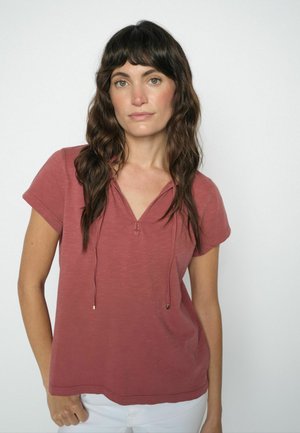 Žena s dlhými vlnitými hnedými vlasmi, oblečená v krátkorm rukáve hrdzavo sfarbenom tričku a bielych nohaviciach, stojaca pred jednoduchým svetlým pozadím.