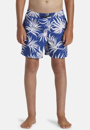 Niño con pantalones cortos de baño azules con patrones de hojas tropicales blancas y rosas, de pie descalzo sobre un fondo blanco.