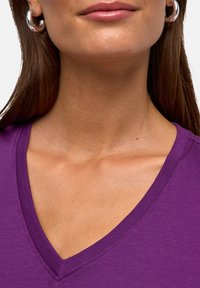 Fioletowa bluzka z dekoltem w serek o gładkiej fakturze, z estetycznym obszyciem, uzupełniona srebrnymi kolczykami-kołami na zbliżeniu.