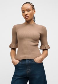 Femme aux tresses cornrows portant un haut beige côtelé à manches courtes avec des détails à volants et un jean bleu foncé, les mains dans les poches.