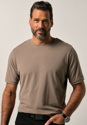 Kurzärmeliges T-Shirt mit Rundhalsausschnitt aus taupefarbenem Stoff, das eine glatte Textur aufweist. Kombiniert mit schwarzen Hosen und einer silbernen Uhr als Detail.