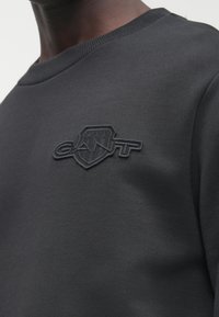 Černá mikina crewneck s texturovanou nášivkou s logem GANT na levé straně hrudi, kterou nosí osoba s tmavou pokožkou.