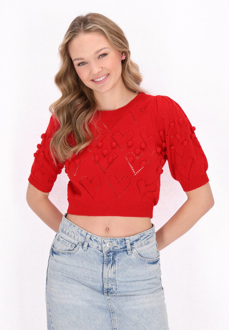 Rød strikket crop top med korte pufærmer, der har hjerteformede udskæringer og strukturerede pomponer, parret med en lys denimnederdel.