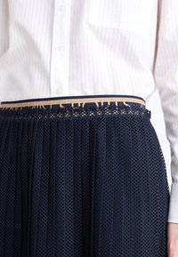 Falda de malla navy con pliegues y una cinturilla decorativa que presenta un logo beige. Combinada con una camisa blanca de botones con rayas.
