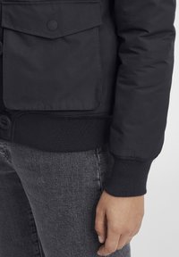 Svart, vadderad jacka med en framficka, ribbade ärmar och en slät yta, kombinerad med grå denimbyxor.