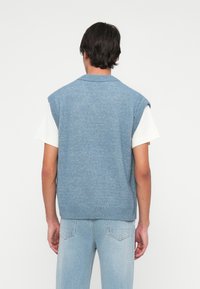 Homem com um camisola de malha sem mangas azul claro por cima de uma camisa de manga curta branca e calças de ganga azul claro, visto de costas sobre um fundo branco.