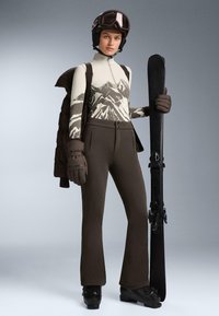 Lyžiarsky oblek s vrchným dielom so zipsom v off-white a šedej farbe s horským vzorom, skombinovaný s hnedými lyžiarskymi nohavicami, rukavicami a lyžiarskou prilbou, drží čierny snowboard.