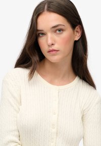 Kremowy kardigan z warkoczykami, z okrągłym dekoltem, z rzędem guzików i fakturowanym wzorem. Miękka, lekka tkanina.