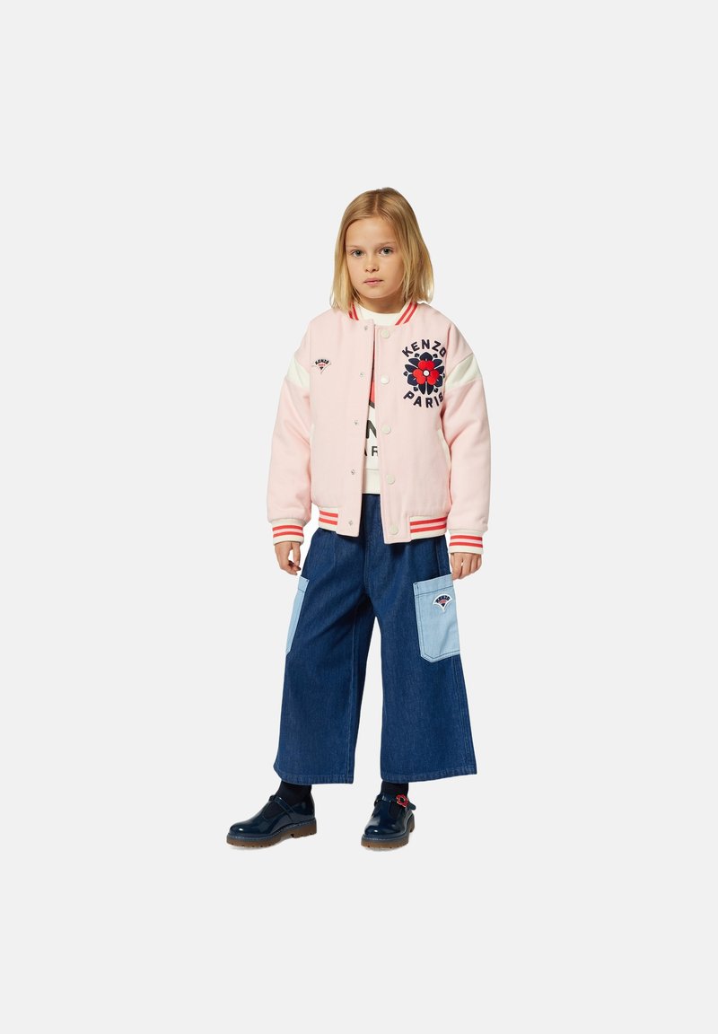 Veste varsity rose avec des manches blanches, des accents rouges, broderies florales, portée sur un t-shirt graphique et un pantalon en denim à jambes larges avec des patchs bleus.