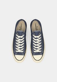 Adidași din pânză navy cu șireturi albe, vârf din cauciuc crem și detalii de cusătură; design clasic low-top, branding proeminent pe interior.