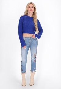 Maglione blu corto con texture a coste, abbinato a jeans chiari strappati con ricami floreali colorati. Stivaletti beige alla caviglia completano il look.