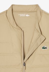 Chaleco acolchado beige con textura suave, cuello alzado, cremallera completa y parche del logo de Lacoste en el lado izquierdo. Diseño ligero y casual.