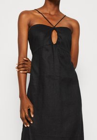 Svart linneklänning med åtsittande liv, spaghettistövlar och ett nyckelhålsutskärning i mitten. Slät textur och flödande silhuett.