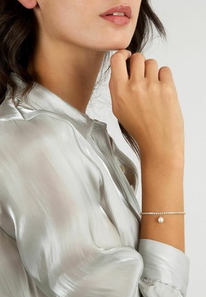 Bracciale d'oro con piccoli cristalli trasparenti e un singolo ciondolo di perla, indossato su un polso, abbinato a una blusa lucida di colore argento chiaro.