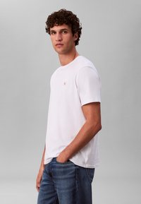 Bijela majica kratkih rukava s okruglim izrezom i malim izvezenim logotipom na prsima. Uparen s plavim trapericama.