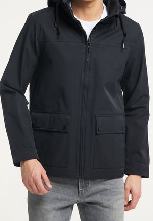 Man wearing black hooded jacket with front zipper and two buttoned flap pockets, paired with light gray jeans.