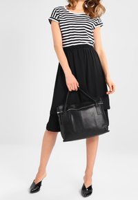 Frau trägt einen schwarzen Rock und ein gestreiftes Hemd, hält eine große schwarze Ledertasche, trägt schwarze spitze Schuhe und Creolen.