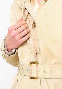 Béžová nepromokavá bunda s dvouřadým zapínáním, páskem s obdélníkovou sponou a texturovanou fleecovou podšívkou na rukávech.