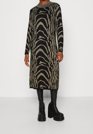 Femme portant une longue robe noire avec des rayures zèbre dorées brillantes et des bottines à talons hauts épaisses noires, debout devant un fond uni.