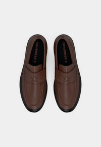 Pantofi din piele maro, fără șireturi, cu o formă rotunjită, având un design cu stea înălțată și o talpă din cauciuc negru. Căptușiți cu interior negru.