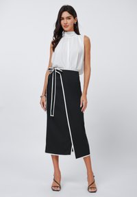 Blusa bianca senza maniche con collo alto, abbinata a una gonna avvolgente nera con un fiocco bianco a contrasto e rifiniture bianche, e uno spacco laterale.