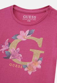 Lyserød T-shirt i økologisk bomuld med et guldglitrende "G"-logo og blomsterdetaljer i pink og grøn. Trykknaplukning ved skulderen.