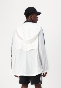 Casaco leve branco com capuz em malha, gola preta e riscas laterais. Usado sobre calções pretos, destacando um design desportivo.