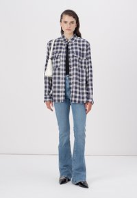 Camisa de botões xadrez em azul marinho e branco, usada sobre uma blusa preta. Jeans flare em azul claro e sapatos pretos de bico fino. Pequena bolsa branca.