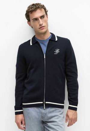Jonge man met krullend haar die een navyblauwe zip-up trui draagt met witte bies en geborduurd logo, lichtblauw overhemd en lichte spijkerbroek.