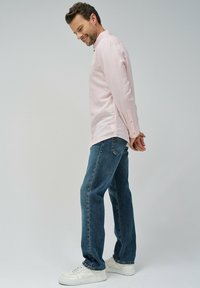 Camisa de manga larga de color rosa claro con puños de botones, combinada con vaqueros azules y zapatillas blancas. El modelo está de pie de lado con las manos detrás de la espalda.