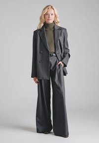 Szary garnitur na miarę z dwurzędową marynarką i spodniami o szerokich nogawkach, noszony na dopasowanym oliwkowozielonym golfie. Gładka faktura.