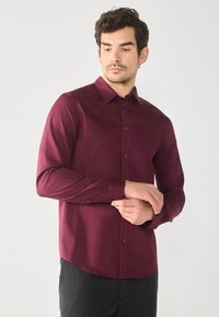Burgunderfarbendes Langarmhemd mit Knopfleiste, Kragen und abgerundetem Saum. Glattes Material mit tailliertem Schnitt und geknöpften Manschetten.