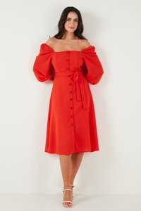 Vrouw die tegen een witte muur staat, gekleed in een rode midi-jurk met blote schouders, pofmouwen, knopen aan de voorkant, een strik in de taille en witte sandalen met hak.