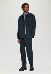 Veste en velours côtelé bleu foncé avec deux poches poitrine, portée sur un col roulé gris et un pantalon assorti, accompagnée de bottes noires à lacets.