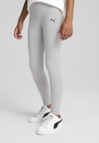 Szürke pamut leggings, amelyek szoros illeszkedésűek, a combon egy kis fekete Puma logóval. Fehér sportcipővel párosítva, fekete részletekkel.