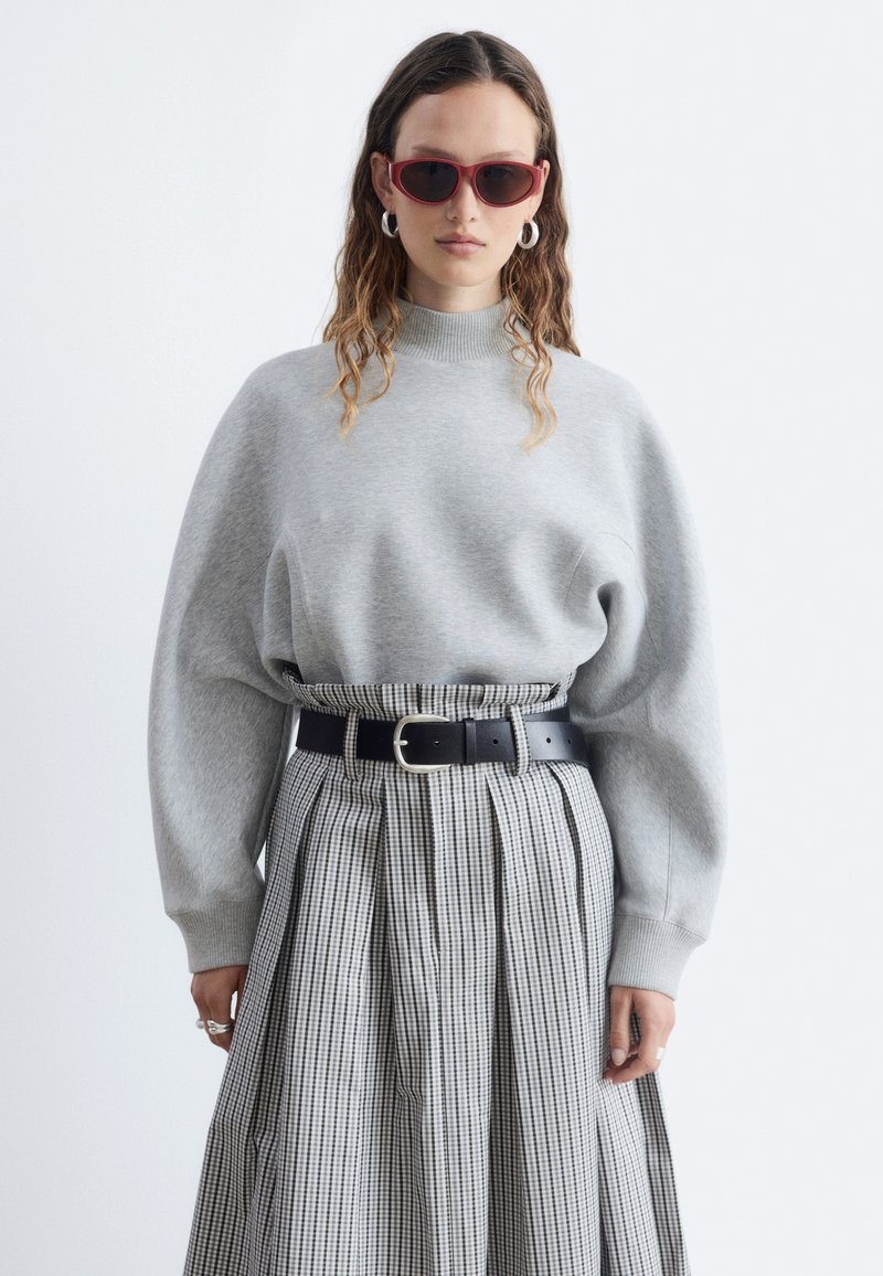 Femme portant des lunettes de soleil rouges, un pull gris clair surdimensionné, une jupe à taille haute à carreaux avec une ceinture noire et des boucles d'oreilles argentées en forme d'anneau.