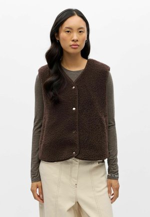 Donna con capelli lunghi e scuri che indossa un gilet in pile marrone scuro sopra una camicia grigia a maniche lunghe e pantaloni crema a gamba larga, in piedi su uno sfondo bianco.