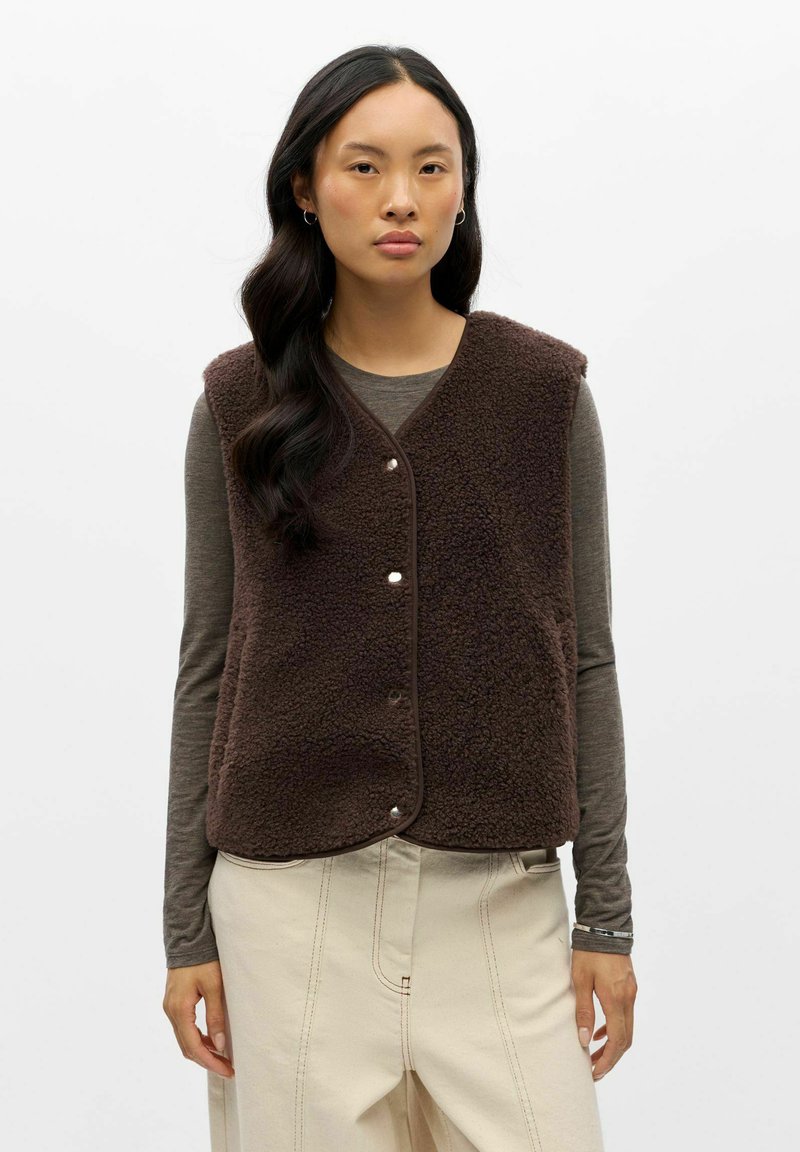 Femme aux longs cheveux foncés portant une veste polaire marron foncé par-dessus une chemise grise à manches longues et un pantalon large crème, debout devant un fond blanc.