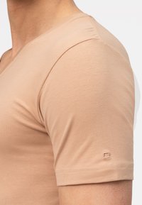 T-shirt in cotone beige chiaro con scollo a V, maniche corte e un discreto dettaglio con logo sulla manica. Texture morbida con design aderente.